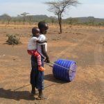young African girl pushing blue water roller with younger brother on her back